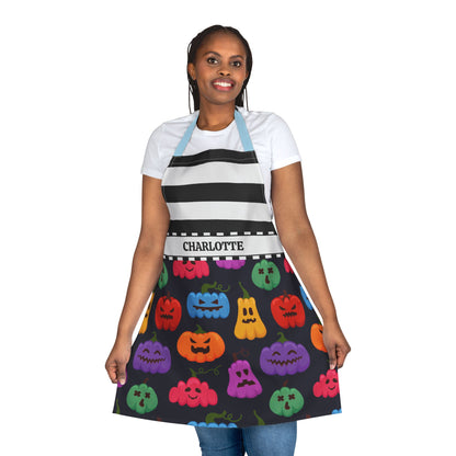 A woman modeling a black kitchen apron with a black and white striped yoke and a colorful Halloween pumpkin print skirt. The apron has a white waistband with the name 'CHARLOTTE' printed on it, and light blue neck and shoulder straps.