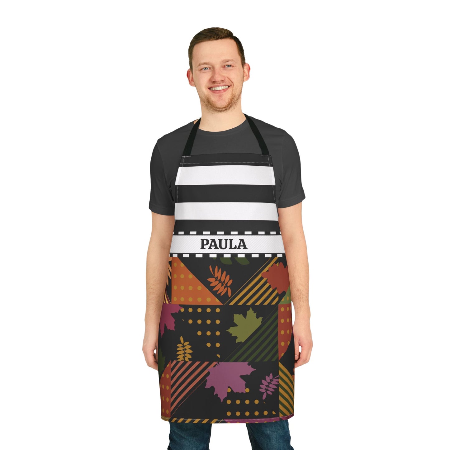 A man wearing a black apron with horizontal white stripes and a colorful autumn-themed pattern on the lower half, featuring the name 'PAULA' in a white waistband