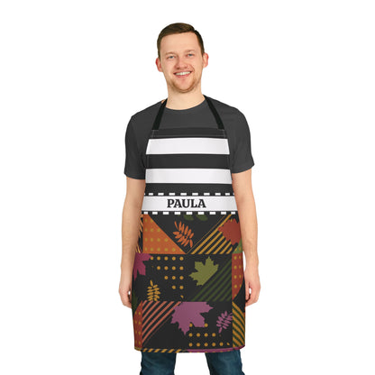 A man wearing a black apron with horizontal white stripes and a colorful autumn-themed pattern on the lower half, featuring the name 'PAULA' in a white waistband