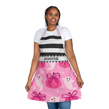 A woman modeling a pink Halloween-themed apron with black and white horizontal stripes on the upper chest, pink bottom section with cartoon ghosts and pink pumpkins with pink bows and a waistband that reads 'JENIFFER', paired with a white t-shirt and blue jeans against a plain white background.