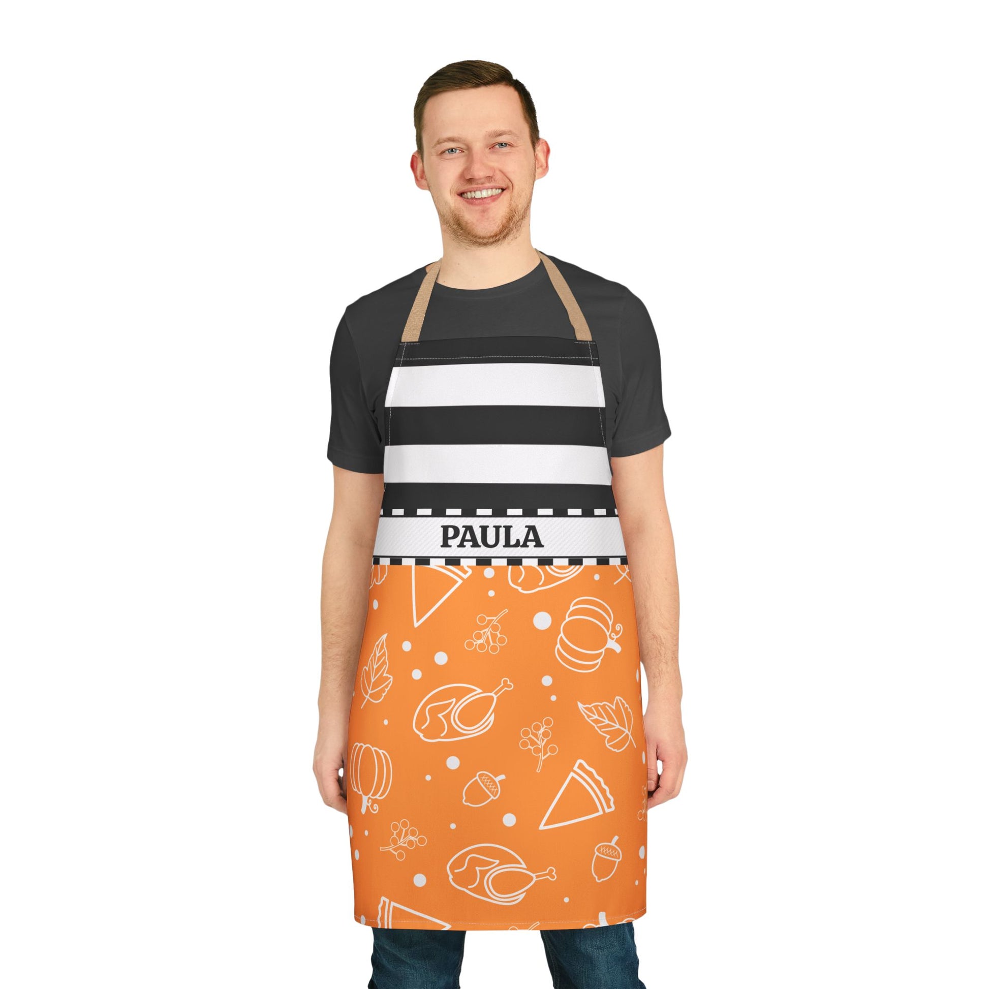 A man wearing a personalized kitchen apron with a black and white striped upper section and an orange lower section with white food illustrations. The apron has the name 'PAULA' printed on a white waistband 