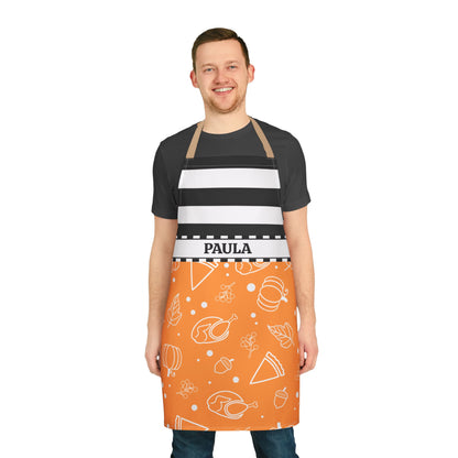 A man wearing a personalized kitchen apron with a black and white striped upper section and an orange lower section with white food illustrations. The apron has the name 'PAULA' printed on a white waistband 