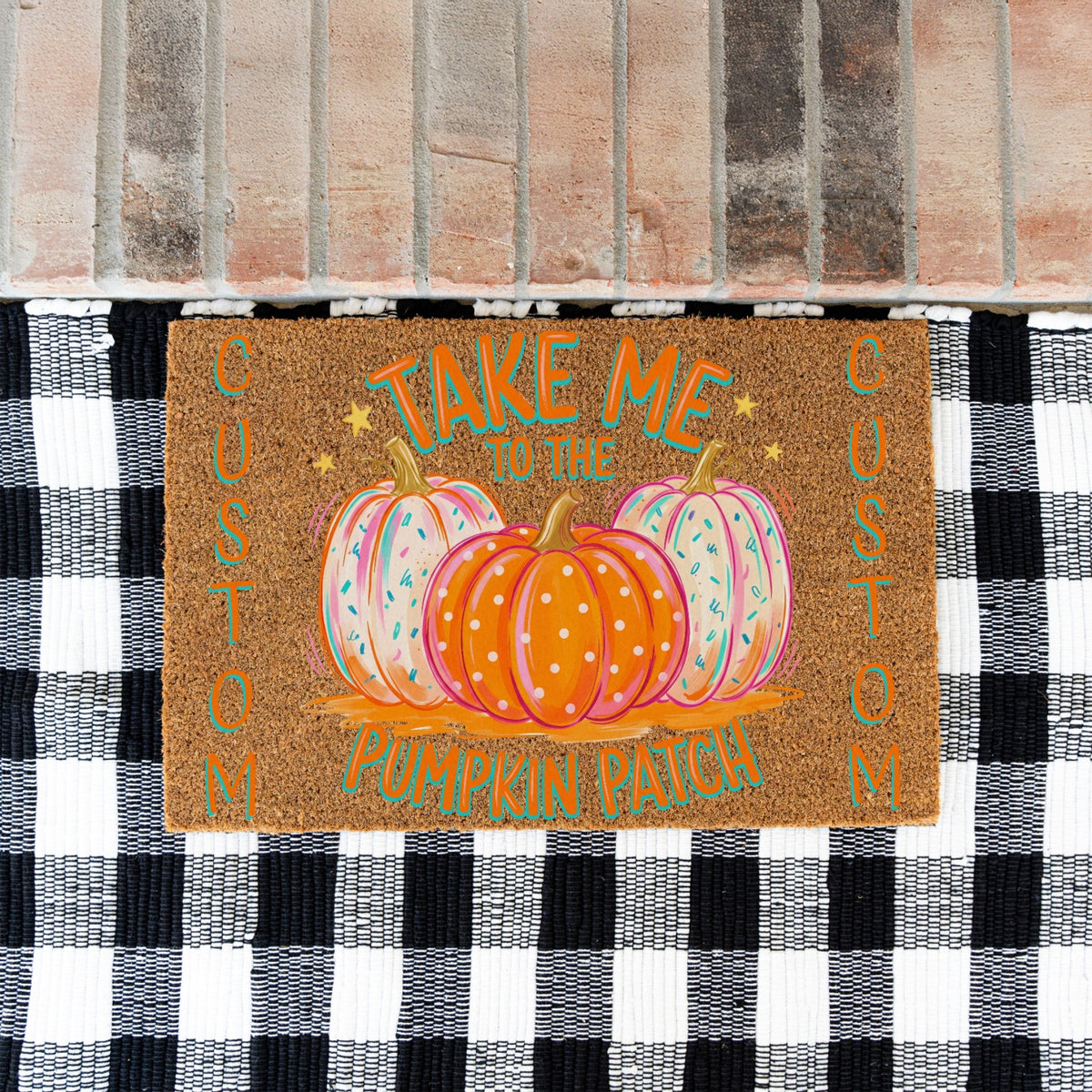 A rectangular brown doormat with a fall harvest design, featuring three pumpkins and the text 'TAKE ME TO THE PUMPKIN PATCH' in teal and orange. The doormat is placed on a black and white checkered rug in front of a brick floor.