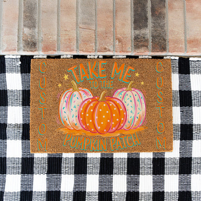 A rectangular brown doormat with a fall harvest design, featuring three pumpkins and the text 'TAKE ME TO THE PUMPKIN PATCH' in teal and orange. The doormat is placed on a black and white checkered rug in front of a brick floor.