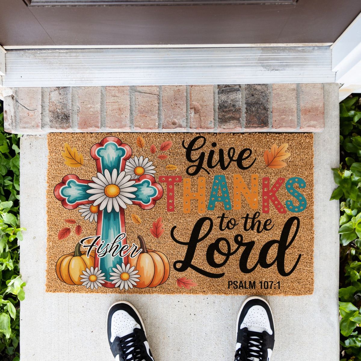 A doormat with a colorful autumn and cross design sits on a concrete porch. The mat features the text 'Give Thanks to the Lord' and custom name 'Fisher' and Psalm 107:1' alongside pumpkins, daisies, and a teal cross.
