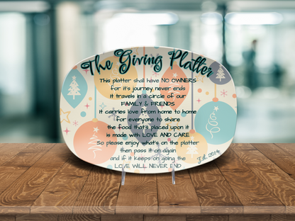 An oval decorative giving platter displayed on small clear stands on a wooden table, with a softly blurred indoor background. The platter has a cream base with pastel blue, peach, and lavender holiday motifs and a large black typographic quote reading 'The Giving Platter, text and custom est year.