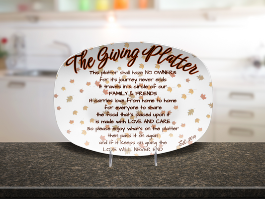 An oval white ceramic giving platter with printed brown text, est year on bottom and scattered autumn leaves, displayed on small clear stands on a dark countertop with a blurred kitchen background.