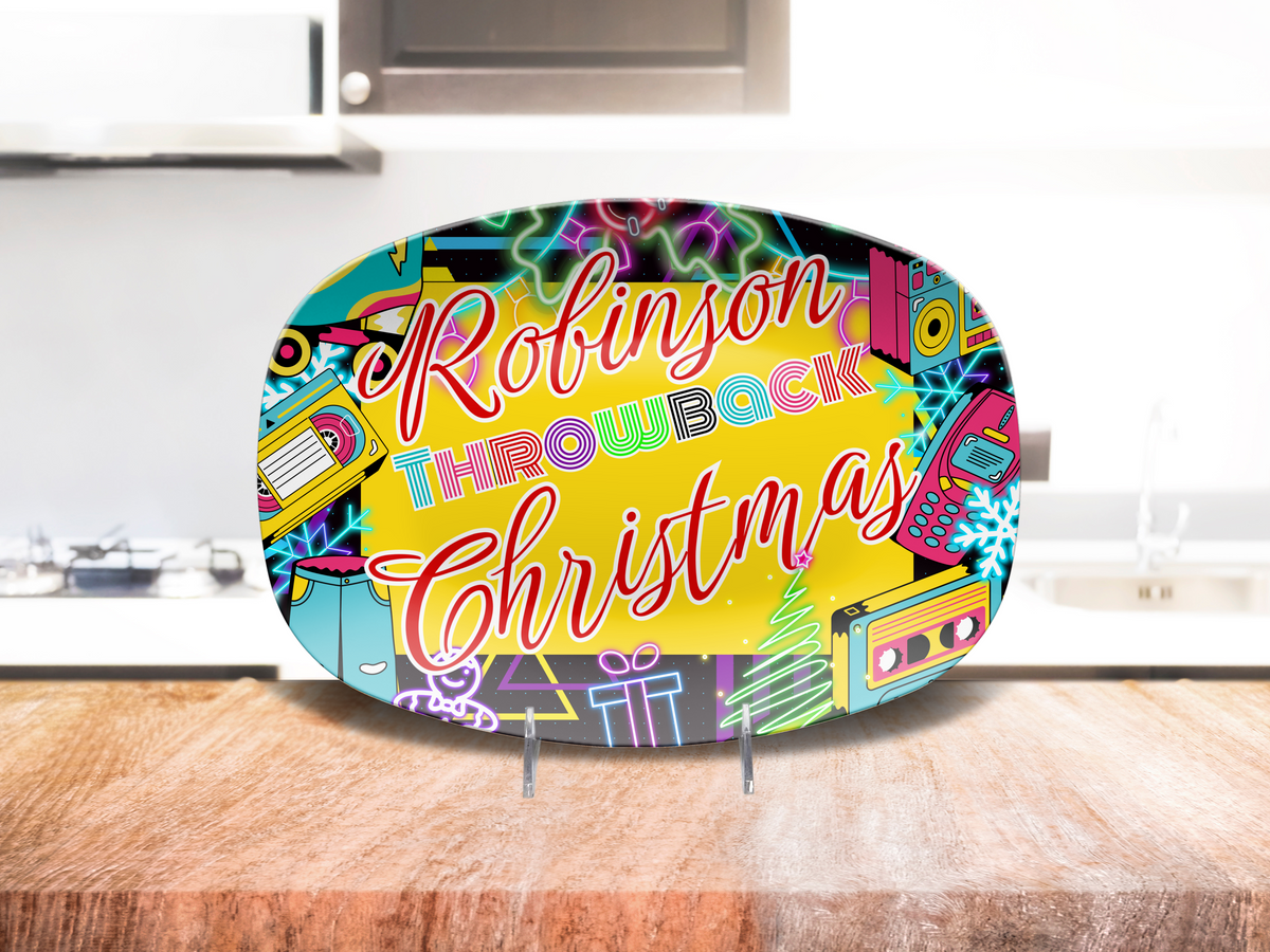 A colorful oval platter on a small stand, featuring the text 'Robinson Throwback Christmas' in stylized lettering over a bright yellow center, surrounded by retro cassette tapes, Christmas tree, snowflake, and other holiday and retro graphics. The sign is photographed standing on a wooden countertop in a kitchen setting.
