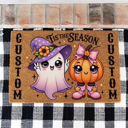 A rectangular coir doormat with a Halloween design, featuring a cute purple ghost in a witch hat and a smiling orange pumpkin with a pink bow. The text 'TIS THE SEASON' is across the top and 'CUSTOM' is printed vertically on both sides.