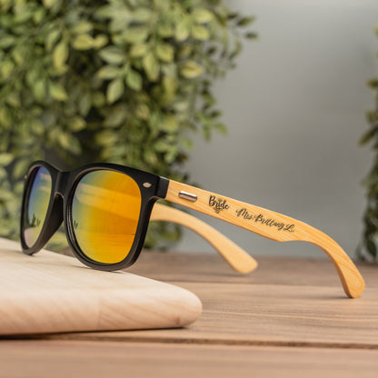 Black-framed sunglasses with mirrored yellow-orange lenses and a natural wood temple engraved with 'Bride  and custom name', resting on a wooden surface with green foliage in the background.