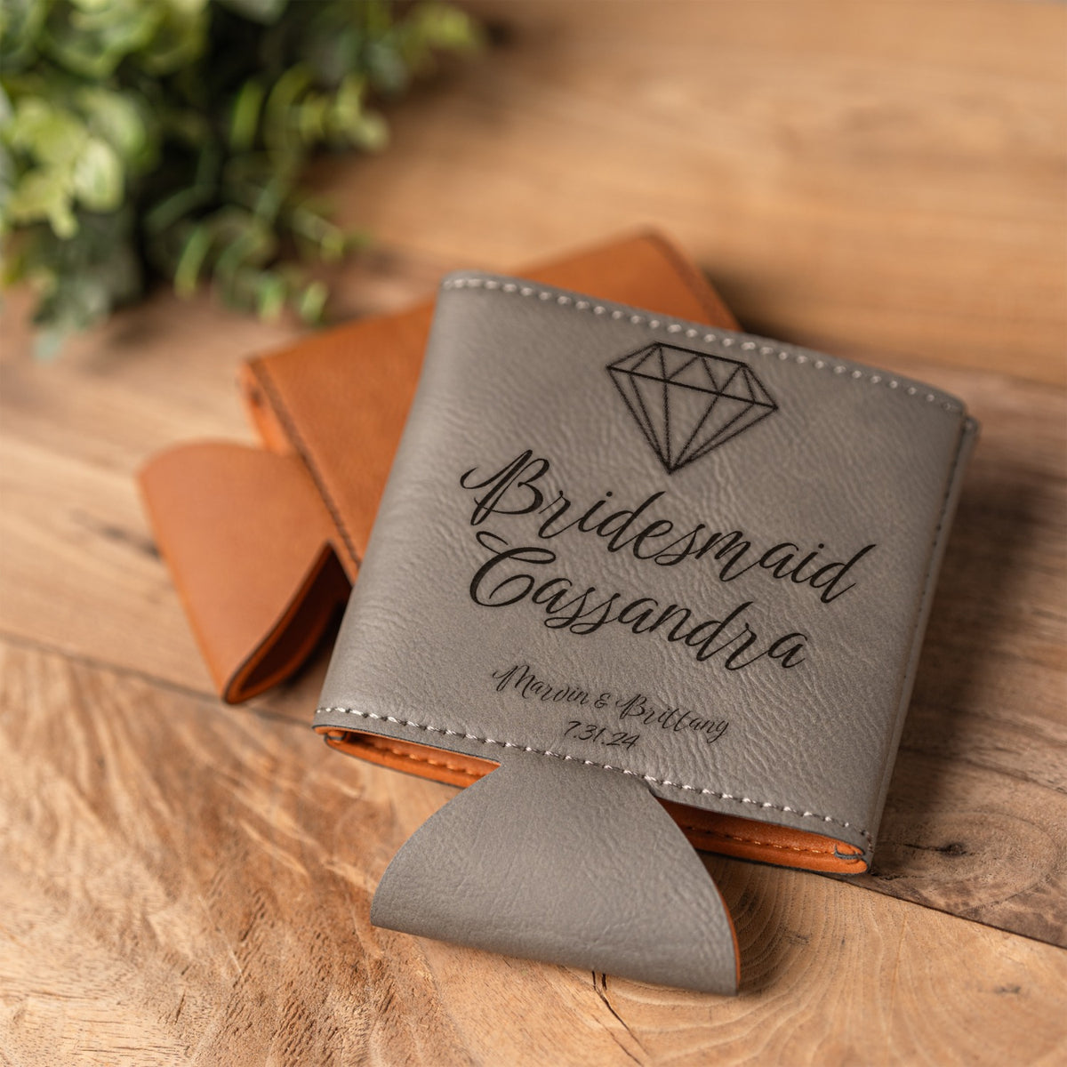 A personalized gray leather can cooler with an engraved diamond icon and the text 'Bridesmaid Cassandra' and 'Marvin & Brittany,' displayed on a wooden surface with a brown leather gift box behind it.