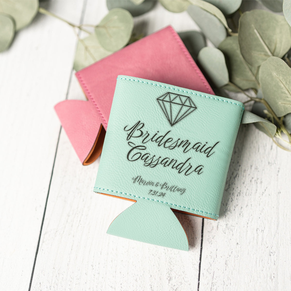 A mint green leather bridesmaid can cooler with a diamond graphic and personalized text, displayed on a white wooden surface with a pink can inserted and eucalyptus leaves in the background.
