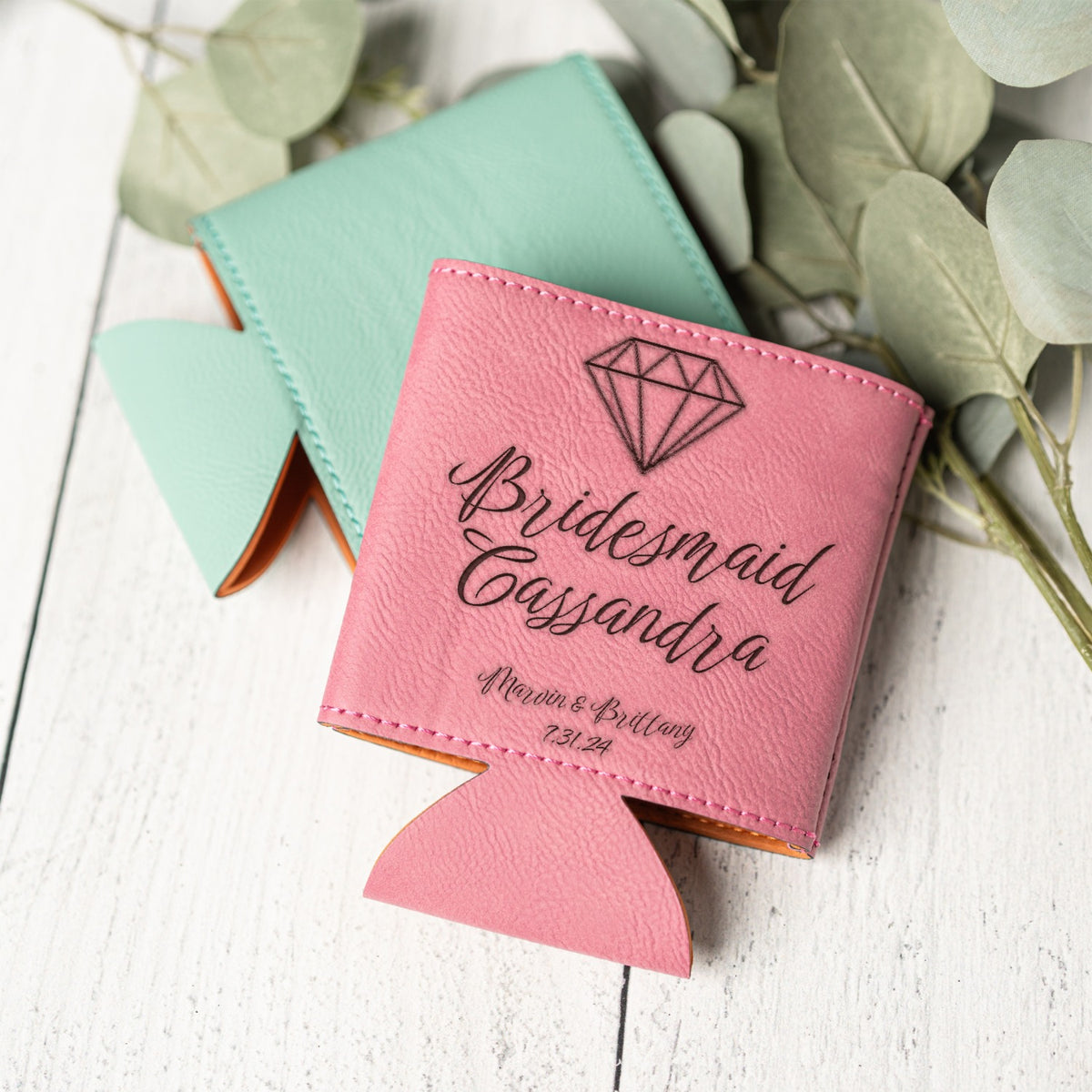 A pink personalized can cooler bottle holder with a diamond graphic and the text 'Bridesmaid Cassandra' and wedding details, shown on a white wood surface with a mint green version behind it and eucalyptus leaves around it.