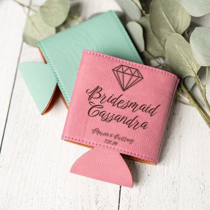 A pink personalized can cooler bottle holder with a diamond graphic and the text 'Bridesmaid Cassandra' and wedding details, shown on a white wood surface with a mint green version behind it and eucalyptus leaves around it.