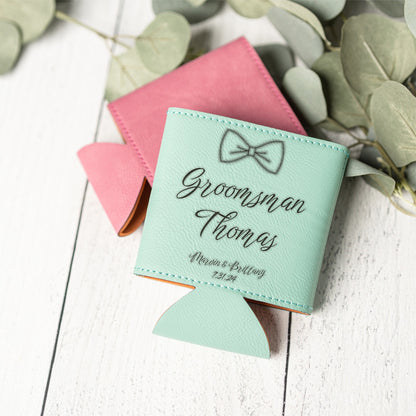 A mint green personalized can cooler with a bow tie graphic and the text 'Groomsman Thomas' on the front, styled on a white wooden surface with a pink can cooler underneath and eucalyptus leaves around it.