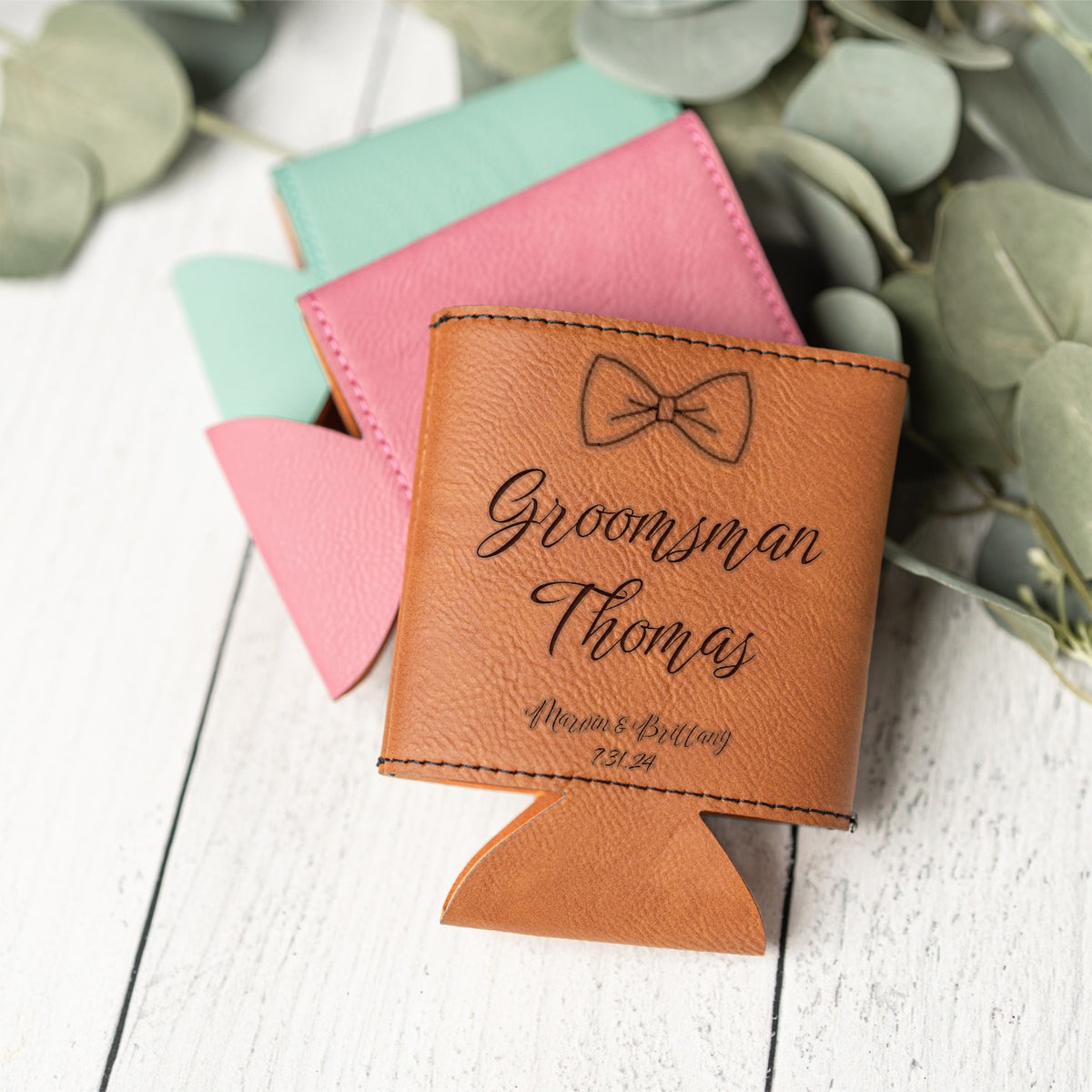 A personalized tan leather can cooler with a bow tie engraving and the name 'Groomsman Thomas', shown on a white wood background with other colored can coolies behind it and eucalyptus leaves around it.