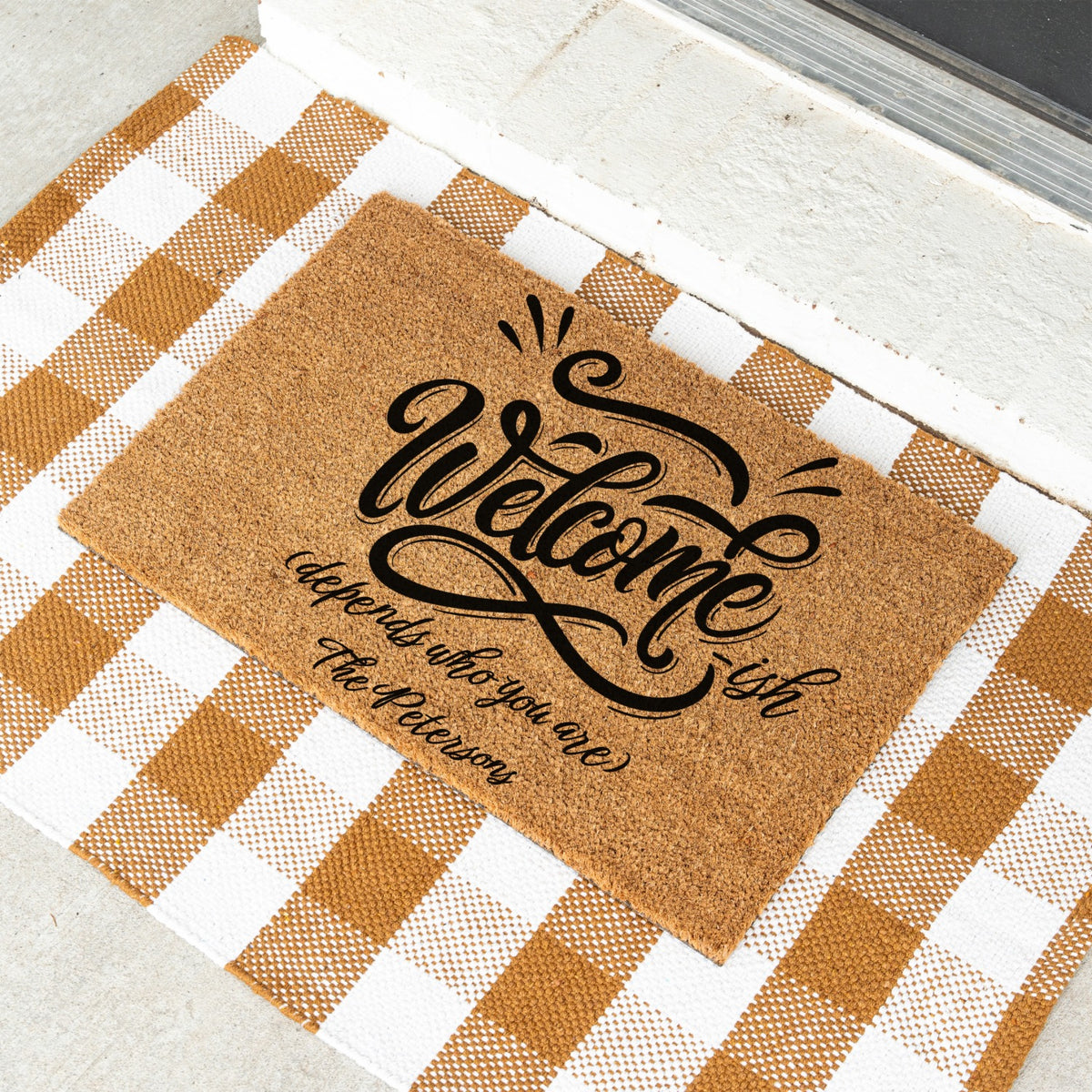 A rectangular doormat with black script reading 'Welcome ish depends who you are, the Petersons' lies on a white and tan gingham check mat in front of a white door threshold.