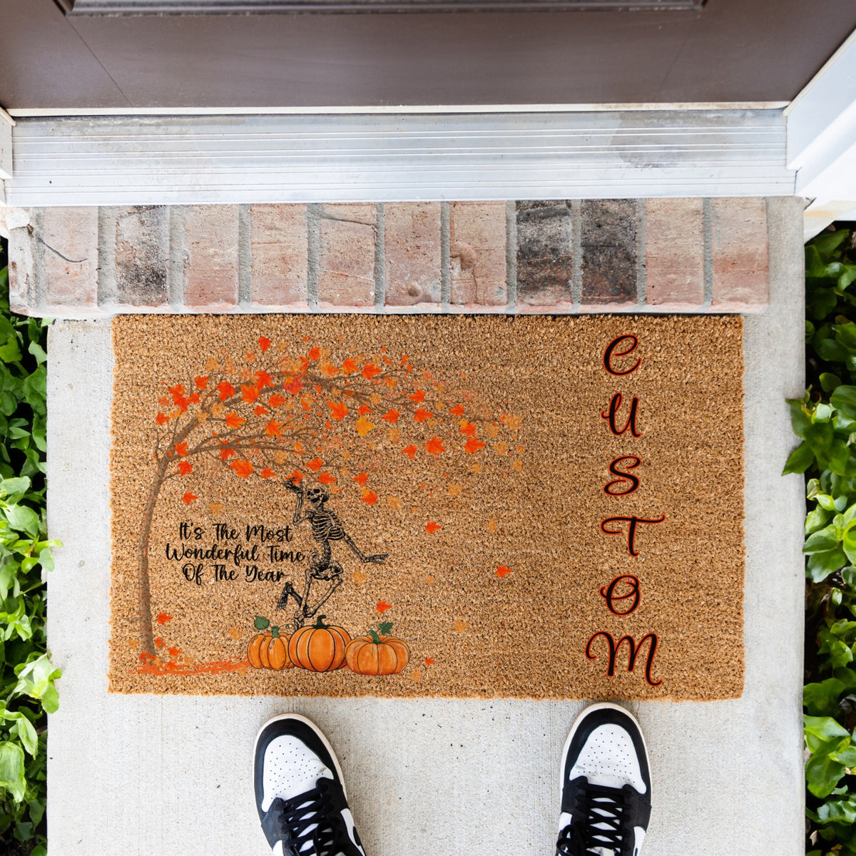 A personalized coir doormat with a skeleton, orange autumn leaves, pumpkins, and the text 'It's The Most Wonderful Time Of The Year' and 'CUSTOM', shown on a concrete porch with a person's shoes visible at the bottom.
