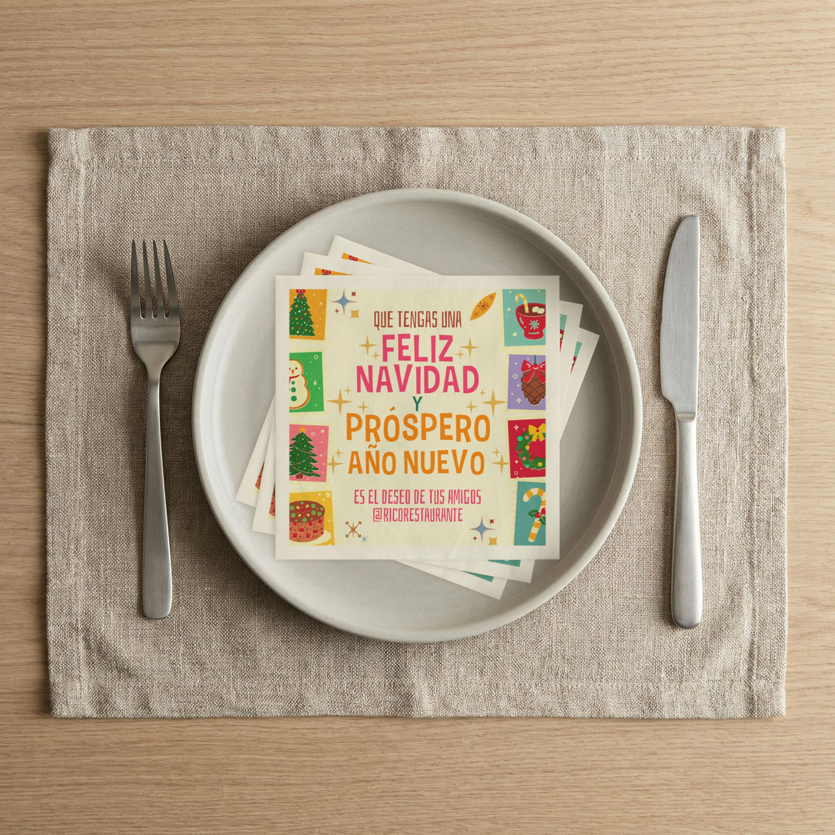 A square napkin with colorful Christmas illustrations and Spanish text reading 'Que tengas una feliz Navidad y prospero Año Nuevo,' styled on a light gray plate with a fork and knife on a beige placemat over a wood table.