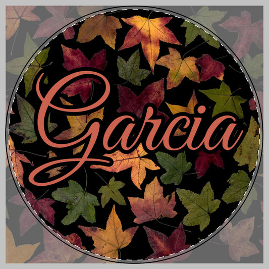 A circular custom cutting board featuring the name 'Garcia' in a coral script font over a background of colorful autumn leaves on a black base. 