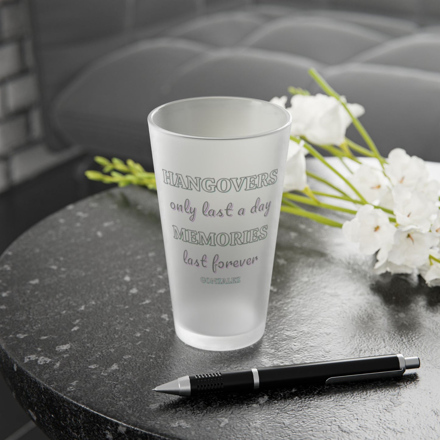 Frosted pint glass that says hangovers only last a day memories last forever and a smaller custom name under that on a dark surface with a pen and white flowers