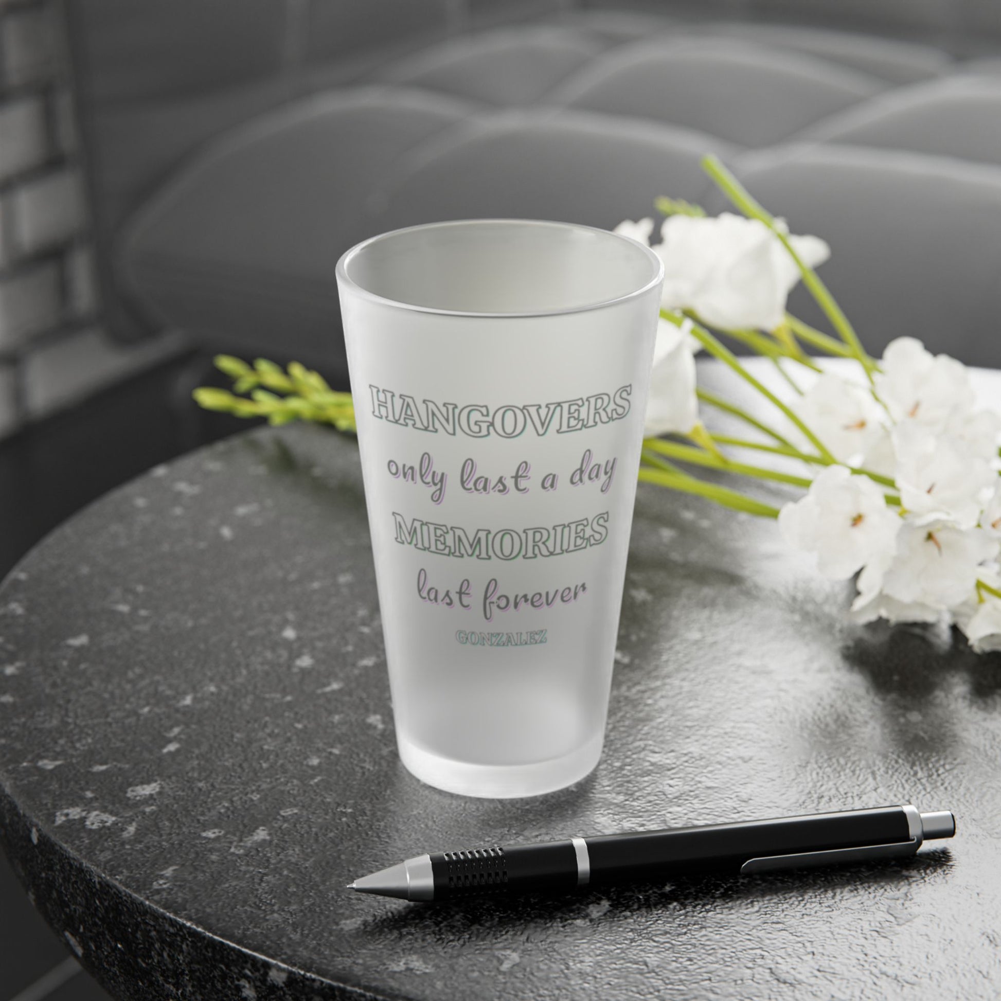 Frosted pint glass that says hangovers only last a day memories last forever and a smaller custom name under that on a dark surface with a pen and white flowers