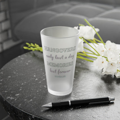 Frosted pint glass that says hangovers only last a day memories last forever and a smaller custom name under that on a dark surface with a pen and white flowers
