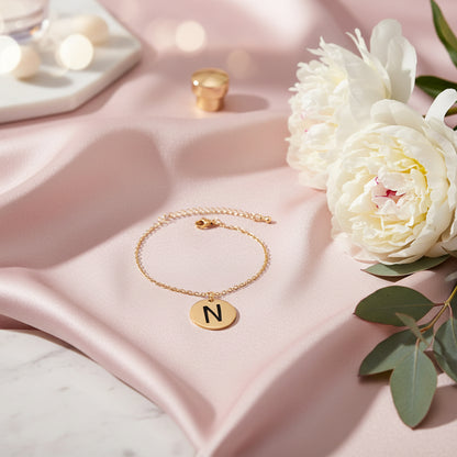Gold bracelet with a round pendant featuring the letter 'N' on a light pink sily material with white roses beside it