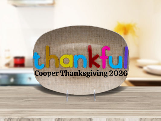 A decorative oval platter with a burlap-textured background, featuring the word 'thankful' in colorful letters and the custom text 'Cooper Thanksgiving 2026' below it. The platter is displayed on a wooden countertop with a blurred kitchen background.